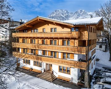A cozy wooden house in the snow with several balconies. In the background, majestic mountains and a clear blue sky are visible.