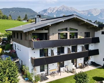 Ein modernes Haus mit mehreren Balkonen, die mit bunten Blumen dekoriert sind. Vor dem Gebäude befinden sich Sitzgelegenheiten und Liegen im Garten.