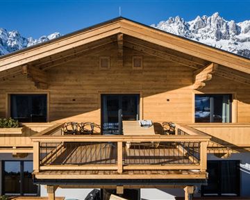 A modern wooden house with a large balcony and a view of the snow-covered mountains. The facade is bright and inviting.