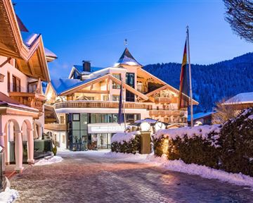 Ein wunderschönes Winterhaus mit schneebedecktem Dach und festlich beleuchteten Fenstern. Im Hintergrund sind schneebedeckte Berge sichtbar.