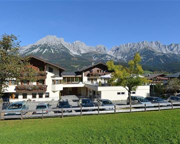 Ein modernes Gebäude mit mehreren Fahrzeugen vor einer schönen Berglandschaft. Im Hintergrund sind hohe Berge und ein klarer blauer Himmel zu sehen.