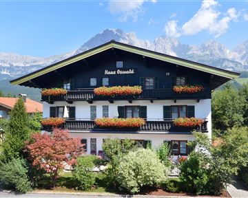 Ein charmantes Gebäude im alpinen Stil mit Balkonen und bunten Blumenkästen. Im Hintergrund sind majestätische Berge und ein klarer blauer Himmel zu sehen.