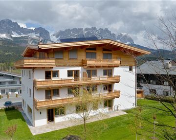 A modern house with wooden cladding and balconies, surrounded by green grass. In the background, impressive mountains and a cloudy sky can be seen.