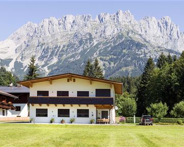 A modern house with a lawn and trees in the foreground. In the background, impressive mountains rise under a clear sky.