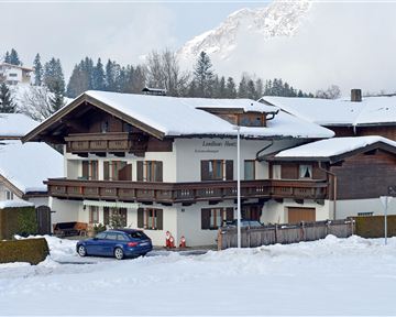 Ein gemütliches Haus im Schnee mit einem blauen Auto davor. Im Hintergrund sind schneebedeckte Berge und Nadelbäume zu sehen.