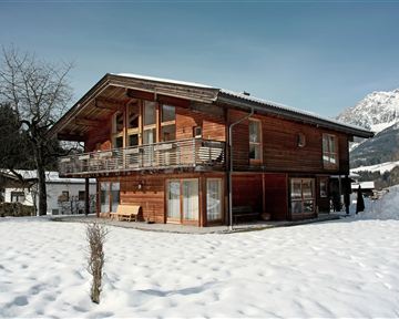 Ein hübsches Holzhaus in einer verschneiten Landschaft. Im Hintergrund sind Berge zu sehen.