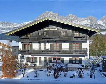 A traditional farmhouse surrounded by snow-capped mountains and trees. The sky is clear and blue, creating a peaceful atmosphere.