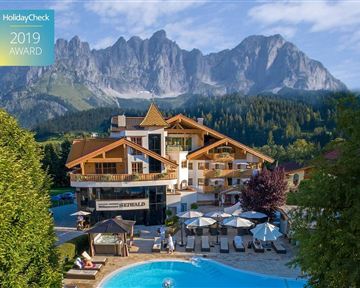 A hotel with a beautiful pool and surrounding loungers. In the background, majestic mountains rise under a clear sky.