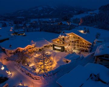A picturesque hotel in a snowy landscape at night. The building is warmly lit and surrounded by snow-covered trees.