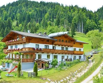 Ein malerisches Holzhaus inmitten der Natur, umgeben von Bäumen und Wiesen. Im Vordergrund befindet sich ein kleiner Spielplatz.