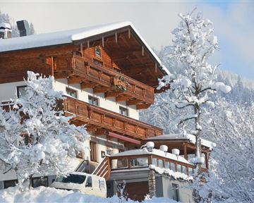 A snow-covered building in alpine style surrounded by winter trees. The sky is clear and the mountains are visible in the background.