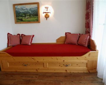 A cozy wooden bed with red bedding and checked pillows. In the background, a landscape painting is hanging.