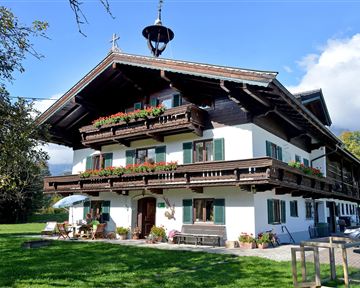 Ein traditionelles alpines Haus mit einem breiten Balkon und blühenden Blumen. Es steht in einer grünen Umgebung unter einem klaren blauen Himmel.