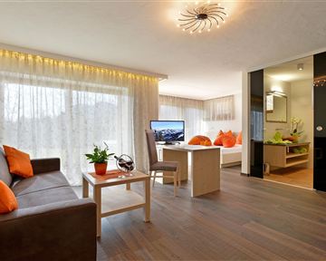 A modern living room with a cozy sofa, a table, and large windows. The orange cushions and plants give the room an inviting atmosphere.