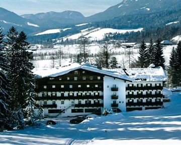 An elegant hotel in the snow with a picturesque mountain panorama in the background. Surrounded by tall, snow-covered fir trees, the winter landscape stretches out.