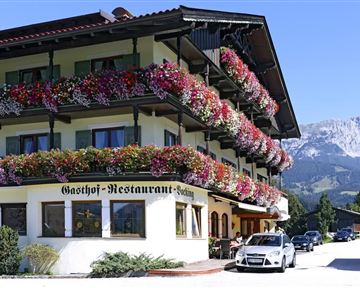 A traditional inn with colorful flower pots. In the background, mountains and a blue sky are visible.