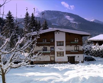Ein gemütliches Chalet im Schnee, umgeben von schneebedeckten Bäumen. Im Hintergrund sind majestätische Berge und ein klarer Himmel zu sehen.