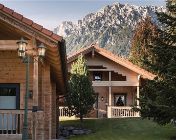 Beautiful wooden cabins in a picturesque landscape with mountains in the background. Surrounded by trees and well-maintained grass.