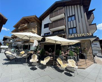 A modern café with cozy seating and sun umbrellas. In the background, you can see a traditional building with wooden cladding.