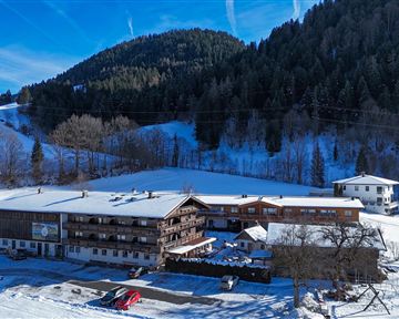 Ein rustikales Hotel in einer schneebedeckten Landschaft. Im Hintergrund sind bewaldete Berge und blauer Himmel zu sehen.