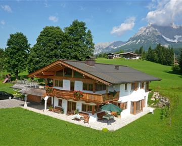 Ein schönes, modernes Haus inmitten einer grünen Wiese. Im Hintergrund sind majestätische Berge und ein blauer Himmel zu sehen.