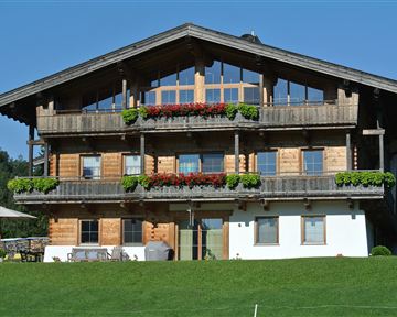 A traditional wooden house with several balconies and colorful flowers. It is located in a green environment under a blue sky.