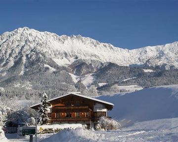 A charming wooden house in a snowy winter landscape. In the background, majestic mountains rise under a clear blue sky.
