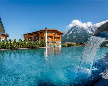 A luxurious pool with clear water and a waterfall. In the background, mountains and a traditional wooden house can be seen.