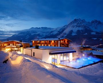 A modern hotel in a snowy mountain landscape at twilight. The outdoor area features a lit pool and snow-covered landscapes.