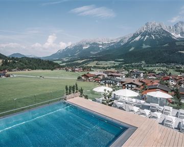 A beautiful view from a pool overlooking a picturesque landscape with mountains and a small village. The surroundings are green and inviting, ideal for relaxation and nature experiences.