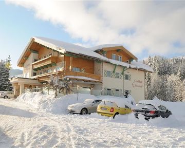 Ein schneesicheres Gebäude inmitten winterlicher Landschaft. Autos sind auf einem verschneiten Parkplatz geparkt, umgeben von schneebedeckten Bäumen.
