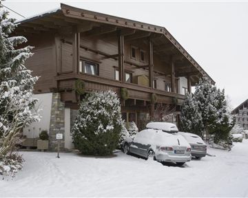 Ein gemütliches Chalet im Winter, umgeben von Schnee. Parkende Autos und schneebedeckte Bäume verleihen der Szene eine ruhige Atmosphäre.