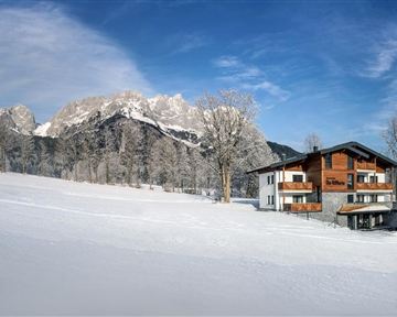 A charming house in a snow-covered landscape with majestic mountains in the background. The trees are frosty and the sky is clear and blue.