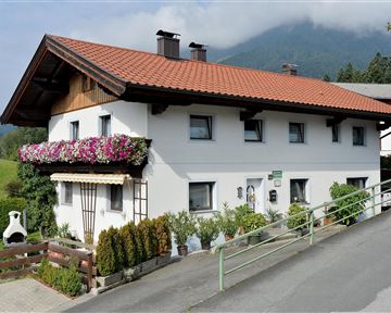 Ein charmantes Haus mit einem roten Ziegeldach und bunten Blumen auf dem Balkon. Im Hintergrund sind sanfte Hügel und eine neblige Berglandschaft zu sehen.