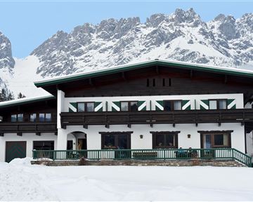 Ein großes Haus im alpinen Stil liegt im Schnee, umgeben von schneebedeckten Bergen. Es hat eine einladende Veranda und große Fenster.