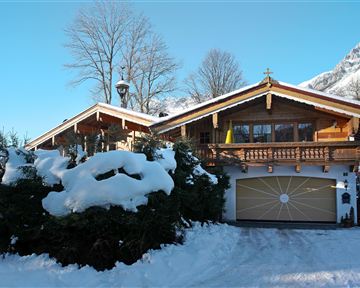 A beautiful wooden house in the snow with a large balcony. Surrounded by snow-covered trees and a clear blue sky.