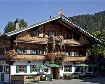 Ein traditionelles Holzhaus mit bunten Blumen und grünen Markisen. Im Hintergrund sind bewaldete Hügel zu sehen.