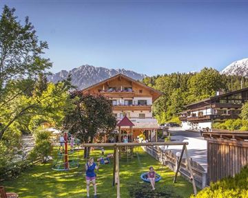 A picturesque mountain village with traditional wooden houses and green meadows. In the foreground, there is a playground and the impressive mountains in the background.