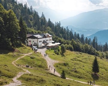 Eine malerische Berglandschaft mit einem Gasthaus und Wanderern auf einem Pfad. Im Hintergrund sind grüne Wälder und Berge zu sehen.