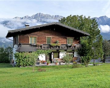 A traditional wooden house surrounded by green areas and mountains. The atmosphere is peaceful and rural.