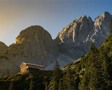 Ein malerisches Bergpanorama mit hohen Felsen und einer Hütte im Vordergrund. Die Sonne scheint sanft auf die Landschaft und die Bäume.