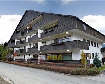 A modern residential building with dark wood design and white walls. The surroundings are green with shrubs and trees.