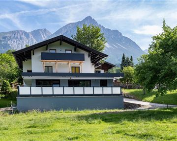 A modern house in front of an impressive mountain landscape. The green meadow and the trees give the scene a tranquil atmosphere.