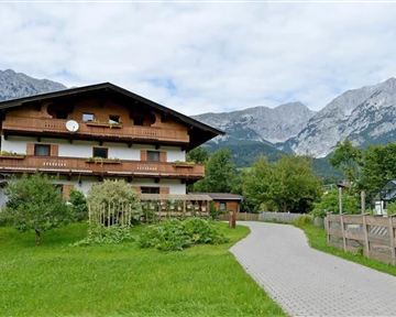 A traditional wooden house surrounded by green landscape and mountains. The sky is cloudy, and a path leads to the front door.