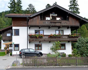 Ein zweigeschossiges Haus im alpinen Stil mit einem Balkon und blühenden Fenstern. Im Vordergrund steht ein Pkw und es gibt einen kleinen Garten mit einem Zaun.