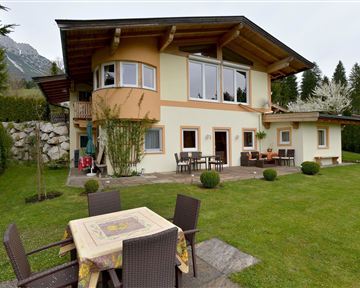 Ein schönes Haus mit einem großen Garten und Terrase. Im Hintergrund sind Bäume und Berge zu sehen.