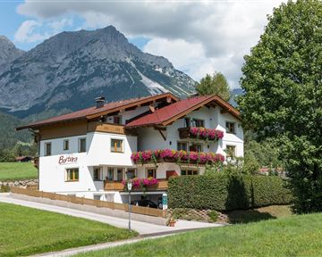 A picturesque house with flower-covered balconies, surrounded by green meadows. In the background, impressive mountains rise beneath a blue sky.