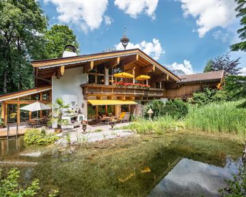 A charming wooden house with a beautiful garden and a small pond. In the background, there are trees and a bright blue sky.