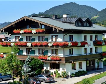 Ein charmantes Hotel mit blühenden Balkonpflanzen und einem malerischen Bergblick. Die Fassade ist freundlich und einladend, umgeben von Grünflächen.