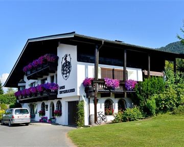 Ein malerisches Gebäude im alpenländischen Stil mit bunten Blumen am Balkon. Umgeben von gepflegtem Grün und einer klaren blauen Himmel.